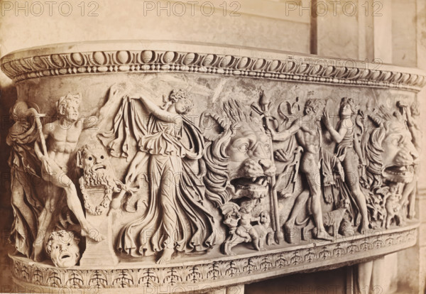 Antique sarcophagus depicting a bacchanal, Museo Vaticano, photograph, photographer: Libreria Spithöver, c. 1850 - c. 1870, pasteboard, paper, albumen print, height 245 mm x width 356 mm, sarcophagus, Vatican City, Vatican Museums, A close-up view of an ornate stone relief frieze showing carved human figures, draped garments, lions with open mouths, small cherub-like children, and theatrical masks. The relief features flowing robes and raised arms, a muscular male figure holding a staff, and a central female figure with a sweeping dress. Two large lion heads with detailed manes face forward, surrounded by smaller decorative elements and wreath-like patterns along the top edge. The stone surface shows variations in tone and texture, with rounded bead-and-egg molding along the upper rim and acanthus-style carving along the lower border.
