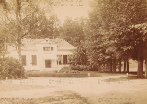 Exterior of a country house on the Zwolseweg in Apeldoorn, photograph, inscription: 'Ant. C. Reelick Photographer Apeldoorn. Special facility for enlargements of any portrait.', photographer: AC Reelick, Apeldoorn, c. 1880 - c. 1900, paper, pasteboard, albumen print, height 97 mm x width 138 mm, rural housing, eg country-house, villa, cottage, façade (of house or building), A two-story light-colored house with a tiled roof is partially obscured by trees and shrubs; the front facade shows shuttered windows and a small central dormer with an oval opening, a doorway with an awning, and a lamp mounted near a ground-level window. Tall leafy trees surround the building, with a grove of darker-trunked trees to the right forming a shaded area, and low hedges and bushes in front. A paved road curves across the foreground with small posts along its edge, and the scene has a soft, faded sepia tone.