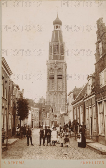 View of Torenstraat in Enkhuizen, with the Zuiderkerk in the background, photograph, inscription: 'Enkhuizen', photographer: Albert Dekema, Enkhuizen, c. 1900, paper, pasteboard, daylight gelatin silver print, height 148 mm x width 97 mm, street, church (exterior), Zuiderkerk (Enkhuizen), A narrow cobblestone street lined with brick buildings on both sides leads toward a tall ornate tower with arched openings and a bulb-shaped dome at the top, dominating the center background. People of various ages stand and walk along the street, including groups of children and adults clustered near the foreground and along the sidewalks, some wearing hats and long coats and a woman holding a basket. Windows and doorways of the adjacent buildings are visible, as are rooftops and a tree on the left side. The scene is captured in a sepia-toned photograph with a clear vertical composition emphasizing the tower.
