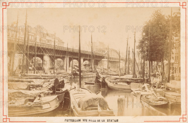 Boats on the Kolk in Rotterdam, Rotterdam vue prize de la station, photograph, Rotterdam, c. 1880 - c. 1900, photographer, paper, pasteboard, albumen print, height 63 mm x width 149 mm, canals, waters (in city), ships (in general), Kolk, A sepia-toned waterfront scene showing numerous moored wooden boats with tall masts filling a narrow canal, a metal arched bridge spanning the water in the background, stone bridge supports and an elevated platform along the left side, multi-story buildings behind the bridge, a row of trees and additional buildings on the right bank, rigging and tarpaulins on many of the boats, and reflections of the boats and masts visible in the water.