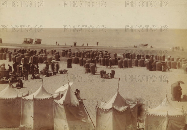 Wicker beach chairs on the beach at Scheveningen, photograph, inscription: 'CP Wollrabe sen, The plate will be kept until reordered.', photographer: Carl Philip Wollrabe, Scheveningen, c. 1870 - c. 1885, paper, pasteboard, albumen print, height 101 mm x width 145 mm, beach (as place of recreation), chair, A wide sandy beach scene with rows of tall, rounded wicker beach chairs scattered across the shore, clusters of small tents with pointed tops and scalloped edges in the foreground, a few figures standing or walking near the waterline, some people seated in and beside the wicker chairs, a pair of wagon-like structures on the wet sand near the water, and a calm sea forming a horizontal backdrop.