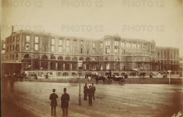 Facade of the Kurhaus in Scheveningen destroyed by fire, photograph, photographer: H. Hille (possibly), Scheveningen, 1886, paper, pasteboard, albumen print, albumendruk, height 96 mm x width 148 mm, ruin of a building, architecture, hotel, hostelry, inn, façade (of house or building), Kurhaus (Scheveningen), A wide, pale-toned photograph shows a large multi-story stone building with rows of arched openings on the lower level and rectangular window openings above, some windows appearing empty. In front of the building is an open paved area with a tall streetlamp near the center. Several men in dark coats and hats stand or gather in small groups across the foreground. A horse-drawn carriage with a driver and a visible horse is positioned near the middle ground in front of the building. Additional carriages and figures are scattered along the facade, and stairways and architectural details are visible on the building's exterior. The scene has an overall muted, sepia appearance.
