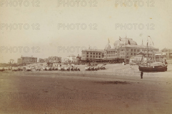 View of the beach at Scheveningen, with the Kurhaus on the right, photograph, inscription: 'CP Wollrabe sen., The plate is kept until reordered.', photographer: Carl Philip Wollrabe, Scheveningen, after 1887 - c. 1900, paper, pasteboard, albumen print, height 97 mm x width 145 mm, beach (as place of recreation), hotel, hostelry, inn, façade (of house or building), Kurhaus (Scheveningen), A wide sandy beach stretches across the foreground with rippled wet sand and scattered darker patches, leading up to a row of small wooden beach huts and horse-drawn caravans lined along the shoreline, each on wheels and clustered near the base of a low sandbank. Behind the huts a large ornate multi-story building with domed towers, arched windows, decorative facades and flags flying from its roof dominates the scene, flanked by several smaller classical and Victorian-style buildings and colonnades. Near the right side of the image a dark-hulled sailing boat with masts and rigging rests on the sand, an...