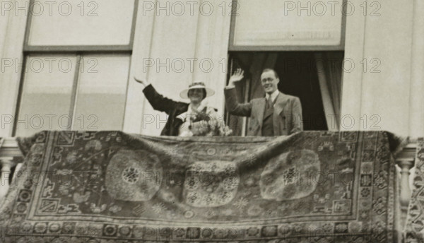 Engagement of Juliana, Queen of the Netherlands, and Bernhard of Lippe-Biesterfeld on the balcony of Noordeinde Palace in The Hague on September 8, 1936, photograph, Netherlands, 1936-09-08, photographer, paper, photography, height 88 mm x width 140 mm, The title mentioned here refers to the highest title obtained by the person portrayed, not necessarily to the title the person portrayed bears in this image., Person, Juliana of the Netherlands, Bernhard (Prince of the Netherlands), The Hague, Noordeinde Palace, Period, A grainy black and white photograph showing a man and a woman standing behind a balcony railing draped with an ornate patterned rug; the woman wears a wide-brimmed hat and a dark coat and holds a bouquet, while the man stands beside her in a suit and tie, both raising one hand in a waving gesture; behind them are tall window frames and curtains, and the rug in the foreground features large circular medallions and geometric border motifs.