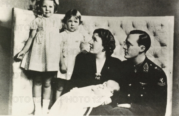 Portrait of Juliana, Queen of the Netherlands, Bernhard van Lippe-Biesterfeld, Beatrix, Irene, Princess of the Netherlands, and Margriet, photograph, Bernhard, Irene and Margriet in Ottawa., Netherlands, 1943 - 1944, photographer, paper, photography, height 89 mm x width 132 mm, The title mentioned here refers to the highest title obtained by the person portrayed, not necessarily to the title the person portrayed bears in this representation., Person, Juliana of the Netherlands, Margriet (Princess of the Netherlands), North America, Ottawa, A black and white photograph shows a seated man in a dark uniform holding a swaddled infant, a seated woman beside him looking toward the man, and two young girls standing behind them on a cushioned backdrop. The standing girl on the left wears a light dress with short sleeves and a bow in her hair, while the other girl wears a lighter dress with a high collar and has short hair. The seated woman has short wavy hair and is wearing a dark outfit with...