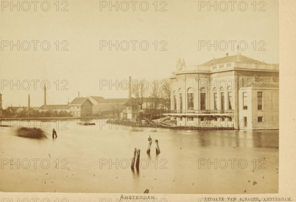 View of the City Theatre on Leidseplein in Amsterdam, seen from the Nassaukade, Amsterdam, cabinet photograph, annotation: 'Nassaukade t, h Leidscheplein', collector's mark: 'VOSMAER', publisher: Andries Jager, 1860 - 1890, Andries Jager, photographer, publisher, paper, pasteboard, albumen print, height 97 mm x width 135 mm, height 107 mm x width 167 mm, canals, waters (in city), theatre (building), Singel, Nassaukade, Schouwburg Leidseplein (1774-1890), A sepia-toned waterfront scene showing a large classical-style building with tall arched windows and a balustraded roofline on the right, adjacent lower structures with rectangular windows, a riverside promenade with railings and small stairways, barren trees behind the buildings, a long low industrial complex with pitched roofs and chimneys stretching along the middle distance, calm water in the foreground with several weathered wooden posts protruding, a small flat boat near the far shore, and blurred motion of a vessel on the left s...