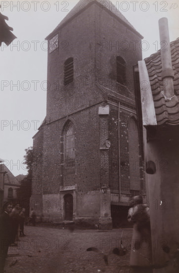 Tower of the Joriskerk in Borculo, photograph, annotation: 'NH Kerk te Borculo', number: '10', annotation: 'NH Kerk, Borculo', Borculo, 1911, photographer, Attributed to, paper, pasteboard, daylight collodion silver print, height 221 mm x width 149 mm, parts of church exterior and annexes: tower, A tall brick church tower with a tiled roof rises from a cobbled street, showing narrow arched windows with slatted openings and a large arched leaded window set into the brickwork, a doorway at the base with a small overhang, and a weathered lower facade; a streetlamp is mounted on the wall near a vertical drainpipe, and adjacent tiled roofs and a chimney are visible to the right; blurred figures of pedestrians appear in the foreground near the base of the tower, and a few trees and rooftops are visible in the background.