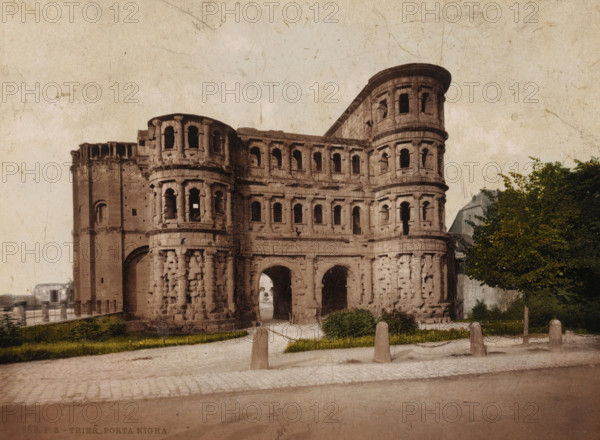 Trier, Porta Nigra, View of the Porta Nigra in Trier, photomechanical print, number: '6950.', cliché maker: Photochrom Zürich, Zürich (city), 1889 - c. 1920, photographer, Rhineland-Palatinate, pasteboard, paper, photochrom, height 188 mm x width 246 mm, city-gate, A large stone gate with three arched openings at the base and multiple tiers of small arched windows rises from a paved area, showing weathered and textured masonry with rounded corner towers; low posts on short chains line a gravel path and grassy patches in front of the structure, while trees with green foliage stand to the right and a faint building rooftop is visible behind them against a pale, slightly stained sky.