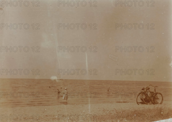 Beach at Valkeveen with people paddling, photograph, inscription: 'N', Valkeveen, c. 1925 - c. 1930, photographer, paper, baryta paper, daylight collodion silver print, height 91 mm x width 119 mm, beach (as place of recreation), A faded sepia beach scene with calm water meeting a sandy shore, small groups of people standing or wading near the water including a person in a dress and hat facing the water, a few distant swimmers, and a cluster of bicycles parked on the right side of the shore.