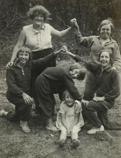 Group portrait of six girls at the Valkeveen campsite, including Koos Ochse, photograph, inscription: 'Ivi Aalkema, Nannie Obbes, Marietje Obbes, Puck Obbes, Ans Robbe, Koos Ockse, The campsite, Valkeveen', Valkeveen, c. 1930 - c. 1940, photographer, paper, baryta paper, gelatin silver print, developed gelatin silver print, height 114 mm x width 90 mm, Person, Koosje Ochse, Ivi Alkema, girl (child between toddler and youth), A group of six girls posed outdoors on bare ground with grass and leafless branches in the background; two standing girls at the back hold hands overhead, one wearing a sleeveless blouse and skirt and the other wearing a long-sleeved top, while four girls in front form a playful shape with two kneeling on either side in dark clothing, one bending forward with hands resting on the head of a seated girl in a light patterned dress, and another kneeling with a smile; footwear ranges from light-colored shoes to sneakers, and the scene is captured in a monochrome photogr...