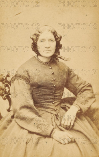 Portrait of a woman from the Marmelstein family, presumably the mother of AF Marmelstein, photograph, carte-de-visite, address: 'A. Greiner, Photograf, Nieuwendijk. L. 87, Amsterdam.', photographer: Albert Greiner, Amsterdam, 1861 - 1874, paper, pasteboard, albumen print, albumendruk, height 105 mm x width 60 mm, historical persons not known by name - BB - woman, A sepia-toned portrait of a seated woman wearing a fitted dress with buttons down the front and decorative trim at the collar and sleeve edges; her hair is parted in the middle and styled in waves with a ribbon tied at the back, and she wears a single earring visible on one ear; one hand rests on her lap while the other is placed on the arm of an ornately carved chair partially visible at the side; the background is plain and lightly textured.