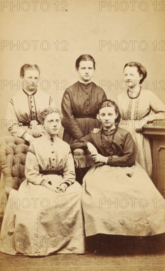 Portrait of a group of young women, one of whom is presumably the mother of AF Marmelstein, photograph, carte-de-visite, annotation: 'Grandma with friends, from Deventer', number: '1869.', photographer: Albertus Josephus van Poorten, Deventer, 1865 - 1869, paper, pasteboard, albumen print, albumendruk, height 101 mm x width 62 mm, anonymous historical persons portrayed in a group, in a group-portrait - BB - woman, Five young women are posed together in a sepia-toned studio photograph. Two women sit in the front on upholstered seats, wearing long skirts and high-collared bodices, one with her hands folded in her lap and the other resting an arm on the chair with a braided necklace and a pendant at her throat. Three women stand behind them, each wearing long dresses with fitted waists and buttoned bodices; the central standing woman has a darker dress and a direct gaze, while the two flanking her wear lighter dresses with decorative trim. An upholstered armchair is visible on the left an...