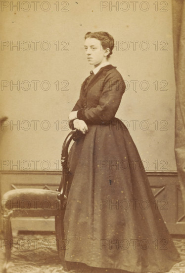 Portrait of an unknown woman from the Marmelstein family, photograph, carte-de-visite, inscription: 'PHOTOGRAPHIE, STAPER & V. CASPEL, Bloem Gracht DD 43, AMSTERDAM.', photographer: Caspel & Stapert, S. van, Amsterdam, 1867 - 1870, paper, pasteboard, albumen print, albumendruk, height 105 mm x width 63 mm, historical persons - BB - woman, A sepia-toned portrait of a young woman standing beside an upholstered wooden chair, wearing a dark, long-sleeved dress with a fitted bodice and a full skirt, her hair styled up and away from her face, hands resting on the back of the chair, a plain backdrop behind her and a patterned floor beneath.