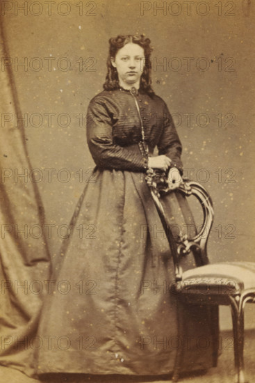 Portrait of an unknown young woman from the Marmelstein family, photograph, carte-de-visite, address: 'Photographisch Etablissement., S. van Caspel & Stapert., Bloemgracht DD 43., Amsterdam', photographer: Caspel & Stapert, S. van, Amsterdam, 1867 - 1870, paper, pasteboard, albumen print, albumendruk, height 100 mm x width 65 mm, historical persons not known by name - BB - woman, adolescent, young woman, maiden, A sepia-toned portrait of a young woman standing and resting her hands on the back of an ornate wooden chair with a cushioned seat, wearing a high-collared dress with a fitted, textured bodice and a full skirt, her hair styled in waves with coils at the top, a curtain draped to one side, and small spots and blemishes visible across the photograph surface.