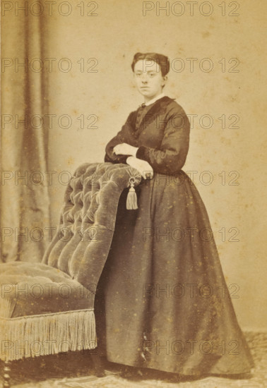 Portrait of an unknown woman from the Marmelstein family, photograph, carte-de-visite, photographer: Caspel & Stapert, S. van, Amsterdam, 1850 - 1900, paper, pasteboard, albumen print, albumendruk, height 106 mm x width 62 mm, historical persons, adult woman, A sepia-toned portrait of a person standing beside a tufted upholstered chair with fringe, the person wearing a long dark dress with a fitted bodice and high collar, hands resting on the back of the chair near a decorative tassel, hair styled back and parted in the middle, and a plain backdrop with a curtain visible on the left.