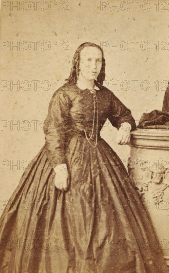 Portrait of an unknown woman from the Marmelstein family, photograph, carte-de-visite, photographer: Jacques Pilartz, Amsterdam, 1850 - 1900, paper, pasteboard, albumen print, albumendruk, height 101 mm x width 62 mm, historical persons, adult woman, A sepia-toned photograph of a woman standing with one hand resting on the top of an ornate pedestal and the other hand hanging at her side, wearing a dark, long-sleeved dress with a fitted bodice and a full, pleated skirt, a light-colored collar visible at the neckline and a long chain hanging from the bodice, her hair parted in the middle and falling in long, smooth waves past her shoulders, the pedestal showing carved decorative faces and scrollwork, and the background plain with light speckling.