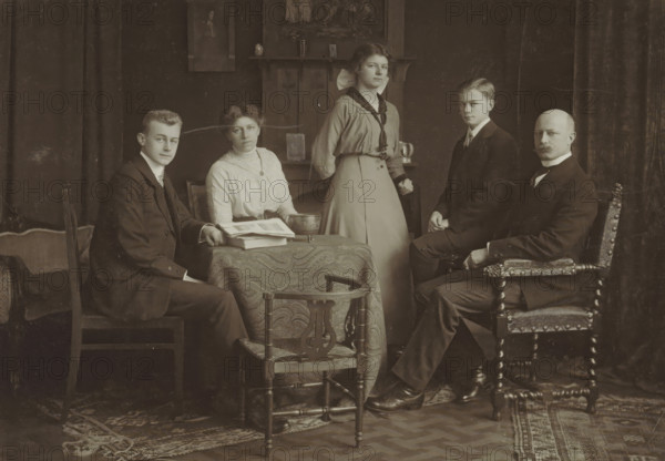 Portrait of a family in a homely environment, probably related to the Marmelstein family, photograph, inscription: 'Familie ...', photographer: Peter Clausing, Haarlem, 1895 - 1920, paper, pasteboard, baryta paper, matte daylight collodion silver print, height 173 mm x width 255 mm, historical persons, adult woman, adult man, A group portrait shows five people posed in a room with dark curtains and a patterned rug on a wooden floor. Three men and two women are present; three are seated and two are standing. The seated man on the left wears a dark suit and sits on a simple wooden chair beside a small table covered with a patterned cloth and stacked books or papers. A seated woman next to him wears a light-colored high-neck blouse and a dark belt, with a shallow bowl on the table beside her. A standing woman behind the table wears a light-colored skirt and a blouse with a dark scarf tied at the neck. A seated young man to the right wears a dark suit and sits on a carved wooden chair, whi...
