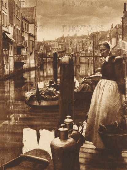 Woman with a basket on a pier in a harbor, photograph, 1850 - 1900, photographer, paper, pasteboard, salted paper print, height 234 mm x width 143 mm, adult woman, harbor, A sepia-toned scene of a canal lined with narrow, tall buildings along the left and distant background, their windows and balconies reflecting in the water; a docked boat filled with goods is moored near a wooden piling in the midground, and a woman in traditional clothing and a headscarf stands on the right near the water's edge with one arm resting on a post and a long skirt falling to her ankles; various jugs and containers sit on the wooden dock in the foreground, and additional small boats and rigging are visible along the canal with rooftops and chimneys forming the skyline under a cloudy sky.