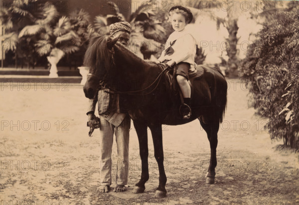 Portrait of an unknown child on a small horse with a servant, cabinet photograph, Illustration of a woman with a map in her hand and a landscape on the verso., photographer: Wah Seng, Makassar, 1905 - 1920, South Sulawesi, Sulawesi, paper, pasteboard, gelatin silver print, height 106 mm x width 166 mm, historical persons not known by name, horse, child between toddler and youth (of undetermined sex), A sepia-toned photograph shows a child seated on a dark horse, wearing a light-colored blouse with a tied scarf and a dark hat, with legs visible and feet in dark shoes resting in stirrups; the horse has a long mane partially covering its face and is fitted with a saddle and reins. Beside the horse stands a barefoot person holding the horse’s reins and wearing striped trousers and a patterned headwrap, with part of their upper body partially obscured by the horse. The background includes large leafy plants and palm fronds, a trimmed hedge on one side, and a flat ground surface in the foreg...