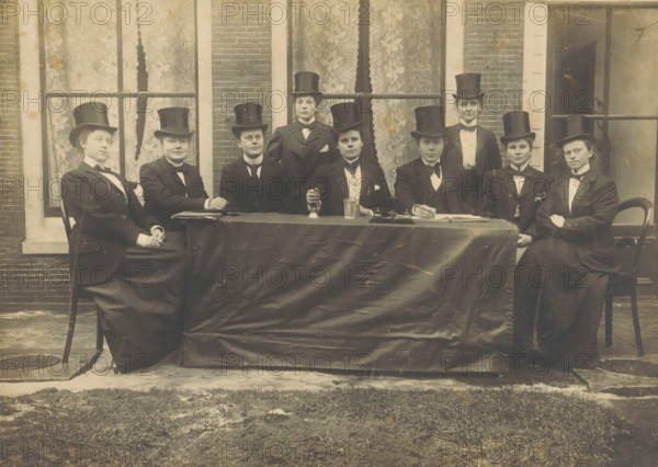Portrait of a group of unknown women with top hats, photograph, 1915 - 1940, photographer, paper, pasteboard, gelatin silver print, developing gelatin silver print, height 128 mm x width 173 mm, head-gear: hat, anonymous historical persons depicted in a group, in a group-portrait, A sepia-toned photograph of a group of people seated and standing behind a long table draped with a dark cloth, all wearing dark suits, white shirts, bow ties, and tall top hats; some are seated in chairs at the table while others stand directly behind, and several faces show neutral expressions; on the table are a bell, a glass, and an inkstand with a pen; the scene is set outdoors in front of a brick building with tall windows covered by patterned curtains, and the foreground shows a paved area and a strip of grass.