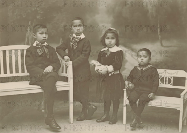 Portrait of four unknown children, probably related to the Marmelstein family, photograph, inscription: '..., Marmelstein-...', photographer: Friedrich Carel Hisgen, Amsterdam, photographer: Julius Heinrich Hisgen, 1888 - c. 1930, paper, pasteboard, baryta paper, gelatin silver print, developing gelatin silver print, height 240 mm x width 319 mm, historical persons, child, Four children are positioned on and between benches in a studio setting. On the left a boy sits with one leg crossed, wearing a dark suit and a large patterned necktie, his hands resting one over the other on his knee. Standing next to him is another boy dressed in a dark coat with a contrasting collar and a spotted scarf tied at the neck, one hand resting on the bench back and the other holding the bench post. Beside him a girl stands in a dark dress with a wide white collar and lace cuffs, holding a small doll in her hands and wearing a bow in her hair. On the right a younger boy sits on a smaller bench wearing a s...