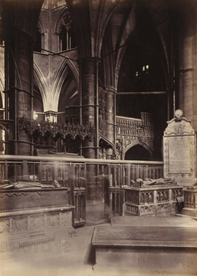 Chapel of St. Edmund at Westminster Abbey, photograph, page, photographer: John Harrington, c. 1864 - in or before 1869, paper, albumen print, height 194 mm x width 139 mm, interior of church, abbey, monastery, convent, Roman Catholic church, parts of church interior: chapel, London, Westminster Abbey, A dimly lit interior of a large stone building with tall vaulted arches and slender columns, ornate carved wooden railings and a decorative screen with intricate tracery extending across the midground, sculpted tombs with recumbent figures on raised platforms in the foreground, a carved monument topped with a rounded finial standing to the right, and layered architectural details including pointed archways and a pulpit or elevated structure partially visible behind the central columns.