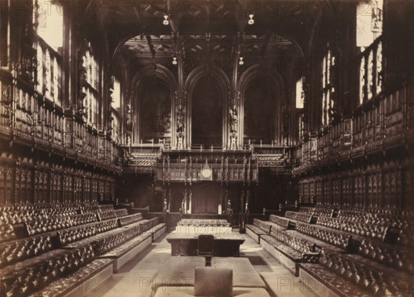 View of the interior of the House of Representatives, photograph, page, photographer: John Harrington, London, c. 1864 - in or before 1869, paper, albumen print, height 140 mm x width 194 mm, palace, interior, representation of a building, Houses of Parliament, A long, ornate hall viewed from one end toward a raised, carved wooden screen and balcony with tall arched openings and decorative columns, flanked on both sides by high, paneled walls with rows of tufted leather benches facing inward, patterned upholstery visible on the seating, a central aisle leading to a table and padded benches in the foreground, tall windows along the side walls admitting bright light, intricate woodwork and sculpted figures incorporated into the balcony and wall panels, and a clock set into the central wooden screen above the entrance.