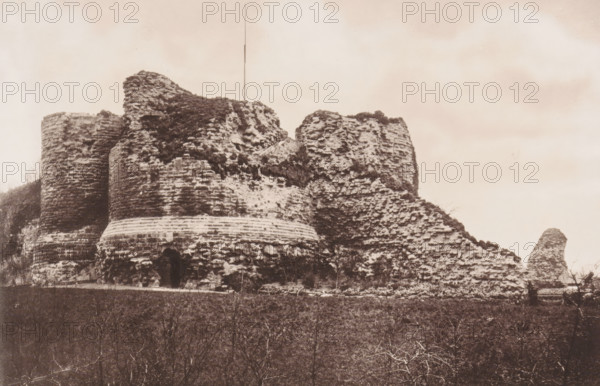 View of the ruins of the round tower of Pontefract castle, Remains of Round Tower. March 31st 1885, page, photomechanical print, Pontefract, cliché maker: Autotype Company, The, 1885-03-31, photographer, paper, collotype, light printing, height 115 mm x width 174 mm, ruin of a building, architecture, castle, A wide sepia-toned photograph of a large ruined stone structure on a hillside, showing rounded towers and crumbling walls with rough, uneven masonry and patches of vegetation growing on and between the stones. The foreground is a darker area with leafless shrubs and low vegetation, and a small entrance or arched opening is visible at the base of one rounded section. A thin vertical pole rises above the highest point of the ruin, and the sky is light and mostly empty above the structure.