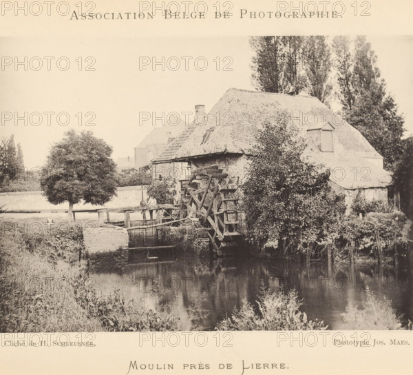 Moulin près de Lierre, View of a watermill, page, photomechanical print, inscription: 'Bulletin de l'Association belge de photographie', maker: Henri Schleusner, maker: Joseph Maes, c. 1885 - in or before 1890, Henri Schleusner, Joseph Maes, paper, collotype, photoprint, height 109 mm x width 155 mm, landscapes with waters, waterscapes, seascapes (in the temperate zone), watermill, A sepia-toned scene shows a riverside building with a steep tiled roof and a large wooden waterwheel partially over the water, vegetation growing on and around the structure, tall trees rising behind it, a leafy tree near a grassy bank, reflections visible in the calm water, a low stone or brick wall along the water's edge, and a person standing on a wooden platform near the wheel.