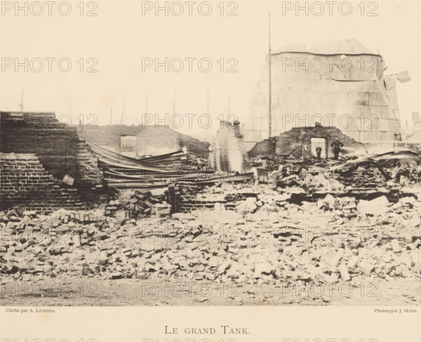 Remains of a large tank from a munitions factory that exploded in Oosterweel near Antwerp in 1889, Le grand tank, page, photomechanical print, number: '18', photographer: Albéric Lunden, Oosterweel, cliché maker: anonymous, Belgium, printer (person): J. Maes (possibly), Antwerp, 1889, printer (person), paper, collotype, photoprint, height 165 mm x width 230 mm, Image on page 18., ruin of a building, architecture, Explosion of a munitions factory in Oosterweel in 1889, A wide scene of rubble and ruined brick walls stretches across the image, with broken bricks and debris scattered densely in the foreground and middle ground. Partially standing brick walls with jagged tops form uneven outlines, and collapsed wooden beams lie atop some of the masonry. In the background a large cylindrical tank with a weathered surface rises behind the ruins, its curved form showing panels and patches. Two small human figures stand near an open doorway in one of the remaining walls, appearing among the des...