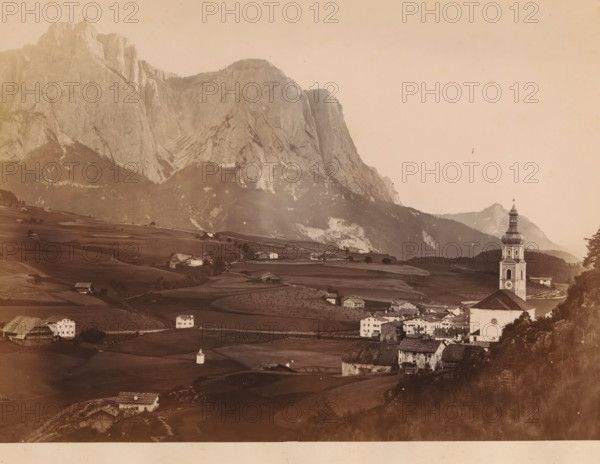 View of Mount Schlern in the Dolomites, Italy, Le Schlern, photograph, page, number: 'N. 14', photographer: Eugène Goblet d'Alviella (possibly), Dolomites, c. 1873 - in or before 1878, Eugène Goblet d'Alviella, paper, albumen print, height 186 mm x width 265 mm, prospect of village, silhouette of village, mountains, Schlern, A sepia-toned landscape showing a village in a valley with rolling fields and scattered farmhouses, a tall church tower with a rounded multi-tiered spire near the right edge, clusters of houses with pitched roofs in the foreground, terraced fields and small isolated buildings across the midground, and a sheer rocky mountain face rising behind the valley and occupying much of the left and central background under a pale sky.