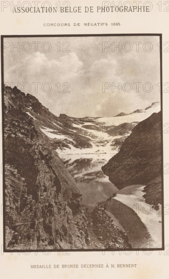 View of a water in the mountains, page, photomechanical print, maker: Angerer & Göschl, C., c. 1880 - in or before 1885, paper, autotypie, height 163 mm x width 122 mm, Print two pages before page 436., landscapes with waters, waterscapes, seascapes (in the temperate zone), mountain-top, peak, A vertically oriented sepia-toned photograph showing a steep rocky mountain valley with jagged cliffs on either side, patches of snow and ice clinging to slopes, a narrow reflective body of water nestled between the rocks, and a sky filled with soft clouds above.