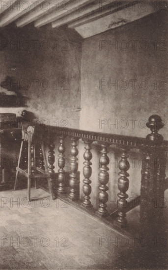 The foundry, l'Atelier des fondeurs, The foundationy, page, photomechanical print, c. 1892 - in or before 1897, paper, collotype, photoprint, height 128 mm x width 178 mm, Person, Museum Plantin-Moretus, workshop interior, Antwerp, A cropped grayscale photograph showing a section of an ornate wooden banister with turned spindles and a rounded newel post, positioned diagonally across the image, adjacent to a textured wall and a tiled floor; part of a stair riser and a portion of a window with blinds are visible at the edges, and a faint shadowed area appears near the lower left.
