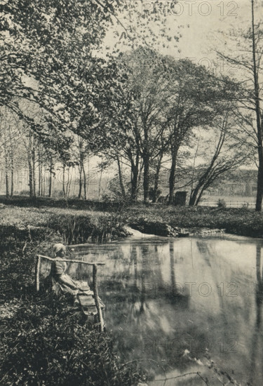 L'Étang de Bel-Air, Près Tours, Woman on the bank of a water feature, page, photomechanical print, inscription: 'Papier &lt, Lustré &gt, . Grosvenor, Chater et Co, 6, rue Favart, Paris.', photographer: Rougé, Tours, cliché maker: Ducourtioux & Huillard, c. 1891 - in or before 1896, Rougé, Ducourtioux & Huillard, photographer, cliché maker, paper, autotype, zincogravure, height 160 mm x width 110 mm, water course (landscape with figures, staffage), A grainy black-and-white scene of a riverside with tall trees along the far bank, their branches and leaves forming a dense canopy that extends across the top of the image, and a calm stretch of water in the foreground reflecting the trees; a small rustic wooden railing and a narrow bench sit at the near edge of the water, where a seated figure in plain clothing and a head covering rests with arms extended to the railing; grasses and low vegetation grow along the riverbank and a small patch of reeds projects into the water, while trunks and t...