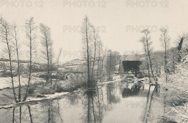 Winter landscape with water feature at Lannion, France, Vieux Moulin sur le Guer, près de Lannion, page, photomechanical print, inscription: 'Papier &lt, Perfection &gt, qualité B. - Grosvenor, Chater et Co, 6, rue Favart, Paris.', photographer: Yves Vallée, Brittany, cliché maker: Berg et Chevalier, c. 1891 - in or before 1896, paper, autotypie, height 120 mm x width 177 mm, winter landscape, landscape symbolizing winter (the four seasons of the year), water course, A riverside winter scene showing bare tall trees along the riverbank, their thin trunks and branches reflected in the calm water, a stone bridge with an arch spanning the river in the midground, a covered wooden structure near the water’s edge, snow on the ground and on some branches, low rolling hills in the background, and the sky occupying the upper portion of the image.