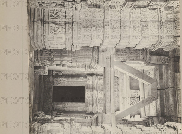 Decorated columns at the great Sasbahu Temple, photograph, page, photographer: KH Mawal, c. 1900, paper, gelatin silver print, developing gelatin silver print, height 150 mm x width 203 mm, temple, shrine, Hinduism, Buddhism, Jainism, column, pillar, architecture, Madhya Pradesh, Central India, Gwalior, A grayscale photograph of a carved stone interior showing a passage framed by intricately detailed columns and beams with repeating floral and geometric reliefs, figurative sculptures, and ornamental panels; the foreground features large angled stone supports crossing the view, while deeper inside a dark rectangular opening contrasts with the lighter carved surfaces; numerous sculpted human and animal figures are integrated into the architectural elements, and layered cornices and pilasters create a sense of depth across the scene.