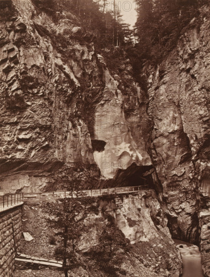 Gorge of Pfeiffers, Switzerland, View of the Aares sky near Meiringen, page, photomechanical print, inscription: 'Europe', photographer: Adolphe Braun & Cie., Meiringen, cliché maker: Woodbury Permanent Photographic Printing Company, c. 1870 - in or before 1875, paper, pasteboard, Woodburytype, Woodburytypie, height 163 mm x width 123 mm, ravine, chasm, abyss, canyon, A sepia-toned photograph showing a steep rocky canyon with textured cliff faces filling most of the frame, jagged rock formations and ledges visible on both sides, a narrow walkway with a railing running along the cliff edge and curving into a dark opening in the rock, scattered small trees and vegetation clinging to crevices, a smooth lighter-toned patch of rock near the center, and a narrow stream flowing at the base of the canyon.