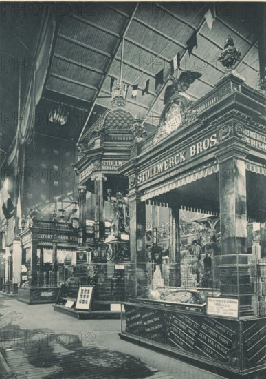 Pavillons en Chocolat (Gebr. Stollwerck, Koeln), Pavilions of the Stollwerck brothers during the 1894 World's Fair in Antwerp, photomechanical print, Part of a cover with 49 plates., inscription: 'Exposition Universelle D'Anvers 1894', photographer: Charles Bernhoeft, Antwerp, Luxembourg (city), c. 1894, printer (person), paper, collotype, photogravure, height 215 mm x width 153 mm, stall, booth (, market), world fair, world exhibition, Wereldtentoonstelling 1894 (Antwerp), Period, 1894, A large indoor scene shows an ornate exhibition space with tall decorative columns, sculpted figures, and elaborate architectural details topped by domes and cresting. A high arched roof with visible rafters spans overhead, and flags or banners hang from above. Carved statues and a winged figure appear atop pedestals, and display cases with angled glass tops contain objects. Potted plants and smaller ornamental elements fill the area, and a patterned wooden floor runs through the foreground. A grand po...