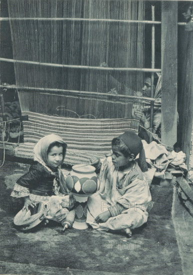 Two unknown Tunisian children in front of an unknown woman weaving a carpet, Tunisia - fabricante de tapis, photomechanical print, Part of a cover with 49 plates., inscription: 'Exposition Universelle D'Anvers 1894', photographer: Charles Bernhoeft, Antwerp, cliché maker: Charles Bernhoeft, Luxembourg (city), c. 1894, Charles Bernhoeft, photographer, cliché maker, printer (person), paper, collotype, photoprint, height 215 mm x width 153 mm, child (at home), Asiatic races and peoples (with NAME), tapestry weaver, gobelin maker, world fair, world exhibition, World Exhibition 1894 (Antwerp), Period, 1894, A woven vertical loom fills the upper background with thin vertical threads and horizontal wooden rods, and a partially completed striped textile hangs from it; in front of the loom two children sit on the ground holding a painted tall ceramic object, the child on the left wearing a headscarf and layered clothing with lace cuffs and holding a small tambourine-like instrument, the child o...