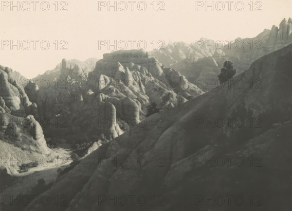 Mountains in Bukhara, Uzbekistan, Alttertiäre Conglomerate an der Oberen Danduschka, page, photomechanical print, inscription: 'Denkschrift d. kais. Akad. d. Wis. math-natural Classe, Bd. LXX.', number: 'Taf. IV.', photographer: A. von Krafft (possibly), Bukhara, cliché maker: Kaiserlich-Königlich Militärgeographisches Institut, c. 1896 - in or before 1901, paper, photoaquatint, height 170 mm x width 220 mm, mountains, A monochrome landscape showing layered rock formations and rugged cliffs receding into the distance under a pale sky, with smooth rounded rock faces and jagged peaks, steep slopes and shadowed valleys, a path or dry streambed winding at the base of the rocks, and a solitary dark tree silhouette on a hillside.