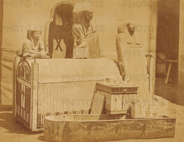 Several sarcophagi in the Boulaq Museum in Cairo, Egypt, photograph, page, photographer: Hippolyte Délié, Cairo (city), photographer: Emile Béchard, c. 1866 - in or before 1871, Ottoman Empire, North Africa, paper, albumen print, height 195 mm x width 251 mm, permanent exhibition, museum, sarcophagus, Boulaq Museum, A sepia-toned scene showing a collection of carved wooden coffins and funerary objects arranged together, including three anthropoid coffins with stylized faces and headdresses, a rectangular chest with vertical panels, a long sarcophagus-like trough in the foreground, small model figures and animal-shaped objects placed atop the chests, and decorative painted panels on the sides of the furniture.