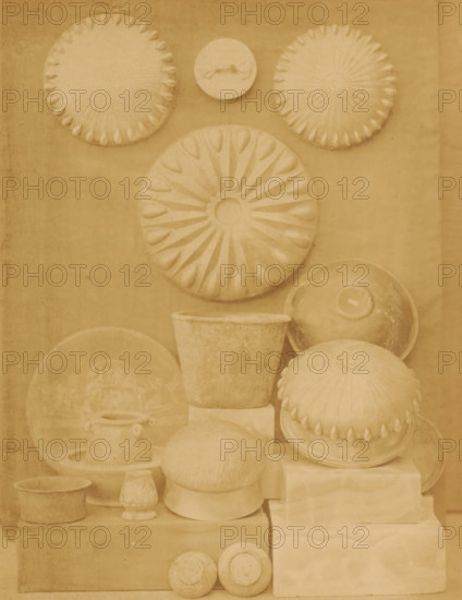 Silver dishes and bowls in the Boulaq Museum in Cairo, Egypt, photograph, page, photographer: Hippolyte Délié, Cairo (city), photographer: Emile Béchard, c. 1866 - in or before 1871, Ottoman Empire, North Africa, paper, albumen print, height 240 mm x width 183 mm, permanent exhibition, museum, bowl, dish, plate, saucer, Boulaq Museum, A vertical arrangement of sculpted round and bowl-shaped objects in a monochrome sepia tone fills the frame, including several large circular discs with radial petal-like patterns, a smaller disc with a handle-like raised feature, domed forms with scalloped edges, shallow bowls and pots, a tapered cylindrical vessel, and stacked rectangular blocks used as supports, all showing textured surfaces and subtle shadows against a plain background.