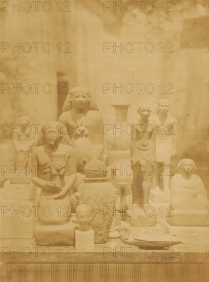 Images from various eras and styles in the Boulaq Museum in Cairo, Egypt, photograph, page, photographer: Hippolyte Délié, Cairo (city), photographer: Emile Béchard, c. 1866 - in or before 1871, Ottoman Empire, North Africa, paper, albumen print, height 232 mm x width 174 mm, piece of sculpture, reproduction of a piece of sculpture, permanent exhibition, museum, Boulaq Museum, A sepia-toned photograph shows a display of carved stone figures and artifacts arranged on tiered platforms, with several human-shaped statues in various seated and standing poses, some wearing headdresses and one holding a small figure; there are rounded vessels, a woven or textured spherical object, and other sculpted items placed among the figures, all set against a plain background with soft shadows.