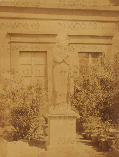 Roman marble statue, found in Tell Mokdam, in the garden of the Boulaq Museum in Cairo, Egypt, photograph, page, photographer: Hippolyte Délié, Cairo (city), photographer: Emile Béchard, c. 1866 - in or before 1871, Ottoman Empire, North Africa, paper, albumen print, height 241 mm x width 184 mm, permanent exhibition, museum, piece of sculpture, reproduction of a piece of sculpture (Roman art), Boulaq Museum, A weathered stone statue of a robed figure stands on a rectangular pedestal in front of a building facade with two recessed window frames; the figure's head is slightly tilted and one arm is raised across the chest while the other rests downward holding a draped garment. Shrubbery and potted plants surround the base of the pedestal and line the ground to the right, with foliage partially obscuring the lower windows. The image has an overall sepia tone and shows signs of age with uneven shading and surface marks across the scene.