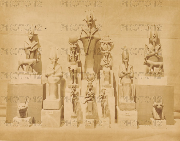 Statues of Osiris and other Egyptian gods in the Boulaq Museum in Cairo, Egypt, Statues of Osiris and other Egyptian idols in the Boulaq Museum in Cairo, photograph, page, photographer: Hippolyte Délié, Cairo (city), photographer: Emile Béchard, c. 1866 - in or before 1871, Ottoman Empire, North Africa, paper, albumen print, height 193 mm x width 252 mm, permanent exhibition, museum, Osiris, god of the underworld, statues, paintings, etc., objects of worship (Egyptian religion), Boulaq Museum, A row of carved figures and animal sculptures arranged on tiered rectangular plinths against a plain wall, each figure carved in a stylized, upright pose with some seated and some standing, many wearing tall headdresses and holding staffs or crossed arms, and two animal sculptures resembling reclining or seated jackal-like heads positioned at the lower corners.
