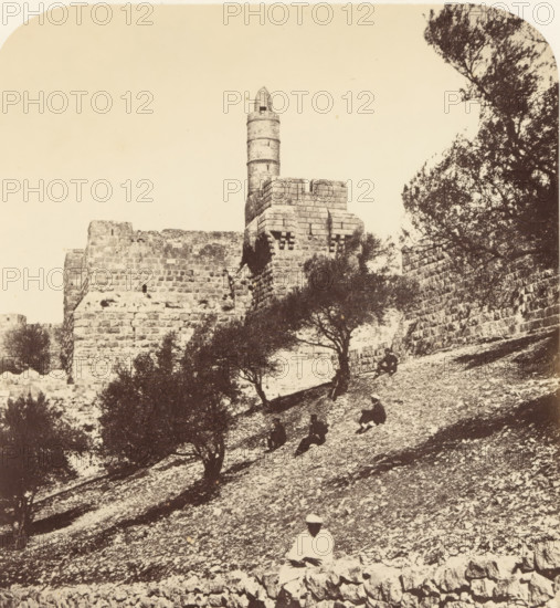 View of the Tower of David in Jerusalem, From the SW outside the Wall of the City, Citadel, photograph, page, photographer: James McDonald, Jerusalem, c. 1860 - in or before 1865, paper, albumen print, height 523 mm x width 340 mm, Photo on page 19., fortified city, Tower of David, A sepia-toned photograph showing a stone fortress wall with a cylindrical tower rising above a crenellated section, framed by trees with dark foliage on the right and scattered trees on a sloping rocky hillside in the foreground, where several seated figures in dark clothing are positioned among the stones and a single figure in lighter clothing sits closer to the bottom edge.