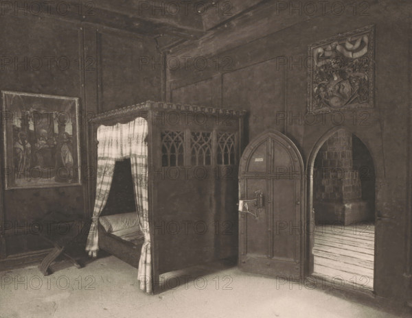 View of a bedroom in the Princely Castle in Merano, Italy, photomechanical print, This print is part of a cover with thirty prints and three text sheets., number: '25.', inscription: 'Déposé.', photographer: Otto Schmidt, Merano, Vienna, printer (person): L. Pisani, publisher: Anton Schroll & Co., c. 1886 - in or before 1891, printer (person), Lower Austria, paper, photoaquatint, height 183 mm x width 236 mm, bedroom, castle - AA - civic architecture: inside, A dim interior scene showing a wooden canopy bed with carved panels and patterned curtains partially drawn to reveal a cushioned mattress, a heavy arched wooden door with metal hinges and a latch, an arched open passageway leading to a space with stacked stone, a framed painting on the wall beside the bed, decorative carved panels above the bed, a carved crest or relief mounted on the wall, a striped wooden floor visible through the arch, and scattered wooden fragments on the floor near the bed.