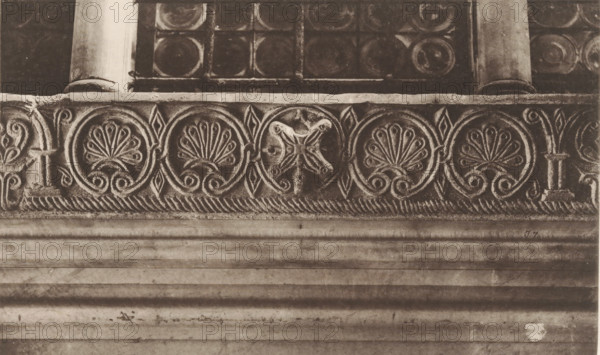 Detail of San Marco in Venice, photomechanical print, This print is part of a cover with 79 prints on 53 pages and 2 text pages., number: 'J.7.', number: 'I.7', number: '215', photographer: Carl Heinrich Jacobi, Venice, photographer: Ferdinando Ongania, c. 1880 - in or before 1885, paper, collotype, photoprint, height 310 mm x width 396 mm, piece of sculpture, reproduction of a piece of sculpture, architectural detail, St. Mark's Basilica, A horizontal stone frieze with repeating carved circular motifs framed by scrollwork sits below a row of small columns and square panels; the carved circles contain fan-shaped floral patterns and one circle shows a cross-like decorative element, all rendered in weathered stone with visible texture and shadowing, above a broad smooth band of layered stone courses that show horizontal grooves, discoloration, and surface marks.