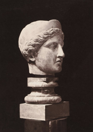 Marble statue of Hera, photograph, page, photographer: Stephen Thompson, c. 1869 - in or before 1874, paper, albumen print, albumendruk, height 239 mm x width 177 mm, piece of sculpture, reproduction of a piece of sculpture, (story of) Juno (Hera), A light-colored marble sculpted head of a person with wavy hair and a smooth cap sits on a polished, rounded pedestal above a rectangular block, all set against a dark background; the face is shown in three-quarter profile with a straight nose, closed lips, and a defined jawline, and the surface of the sculpture shows subtle wear and mottled texture.
