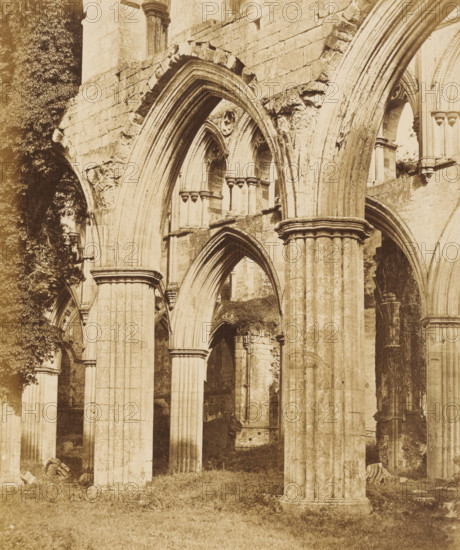 Interior of the choir of the Fountains Abbey near Ripon, photograph, page, photographer: Joseph Cundall, Fountains Abbey, photographer: Philip Henry Delamotte, c. 1851 - in or before 1856, Yorkshire, Studley Royal Park, paper, albumen print, height 280 mm x width 232 mm, ruin of church, monastery, parts of church interior: choir, A sepia-toned photograph of large stone arches and columns from a ruined building, showing rounded and pointed archways with fluted pillars and carved capitals, weathered stone blocks and broken masonry, patches of stone rubble and fragments at the base, grass covering the ground, and a vertical mass of foliage climbing some of the stonework.